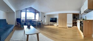 A modern living room with a sofa, dining table, and chairs, a kitchen area with cabinets and appliances, and a large window providing natural light. - Ferienwohnung Nr. 4, für 1 bis 2 Personen, 46 qm, mit Dusche/WC, Wohn-/Schlafraum getrennt (Schönau am Königssee)