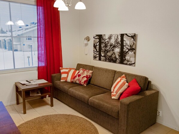 Una sala de estar con un sofá marrón, almohadas rojas y blancas, una mesita de madera y un ventanal con cortinas rojas.