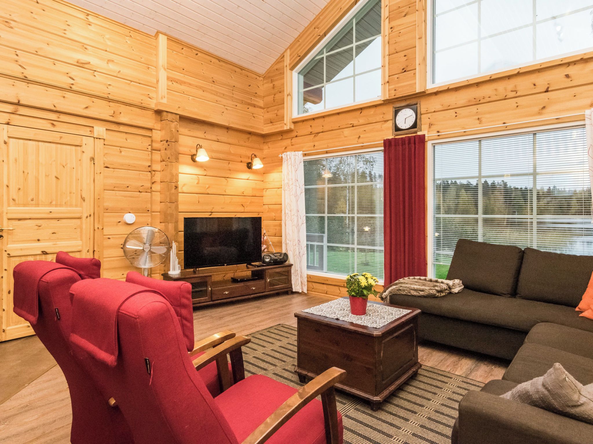 A cozy log cabin interior with a television, red armchairs, a sofa, and a large window with curtains.