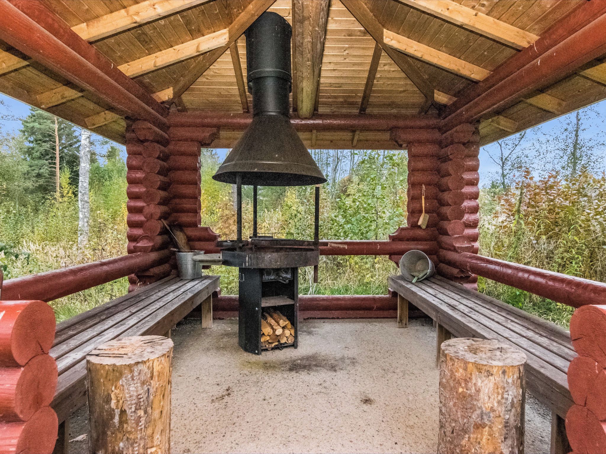 A rustic wooden cabin with a central wood stove, benches, and a view of the surrounding forest.