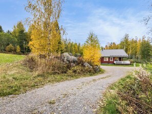 Ein Kiesweg führt zu einer roten Holzhütte mit weißem Veranda, umgeben von Herbstlaub und einem Wald.