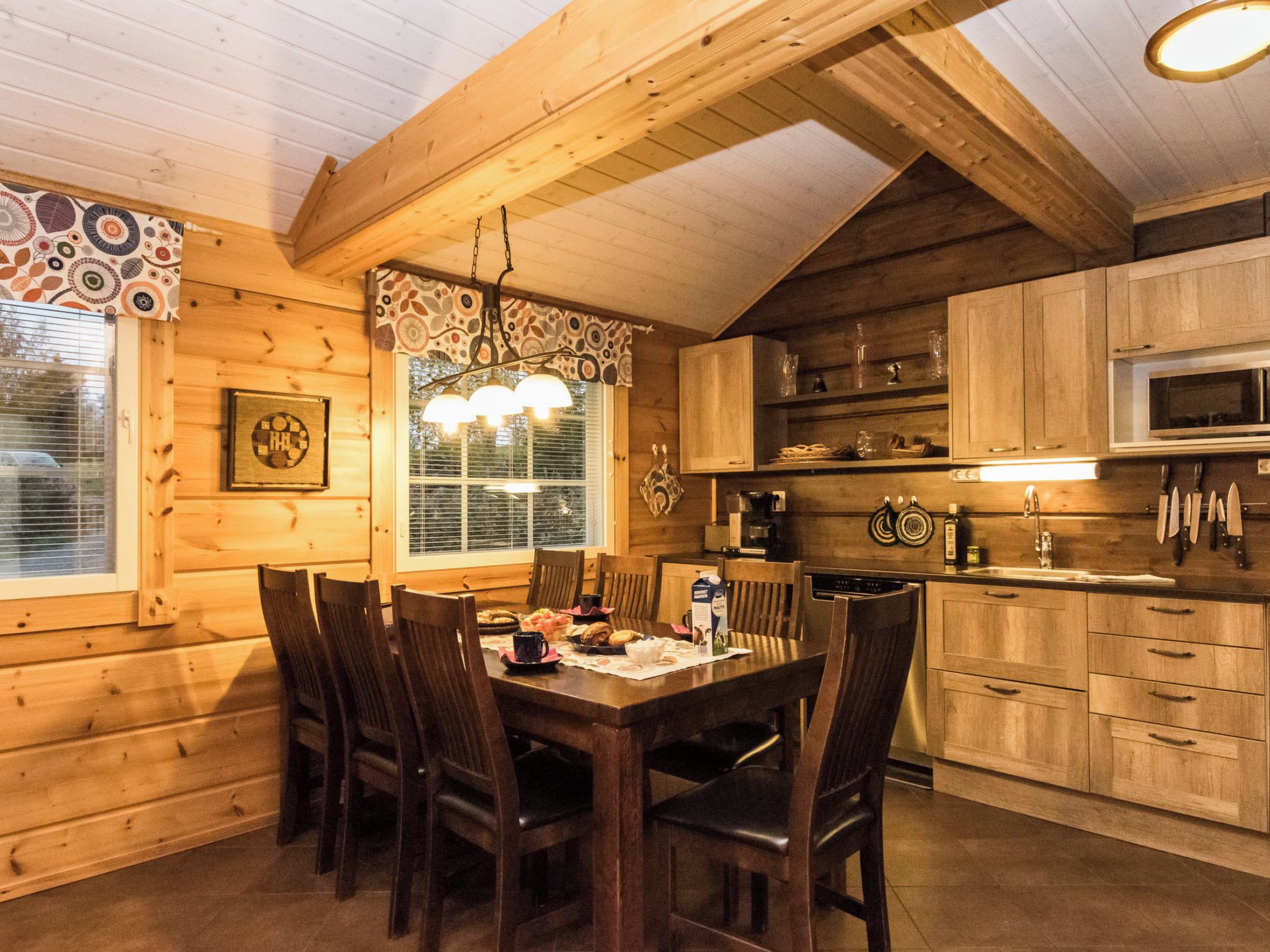 A wooden interior with a dining table set for four, kitchen cabinets, a microwave, and a hanging light fixture.