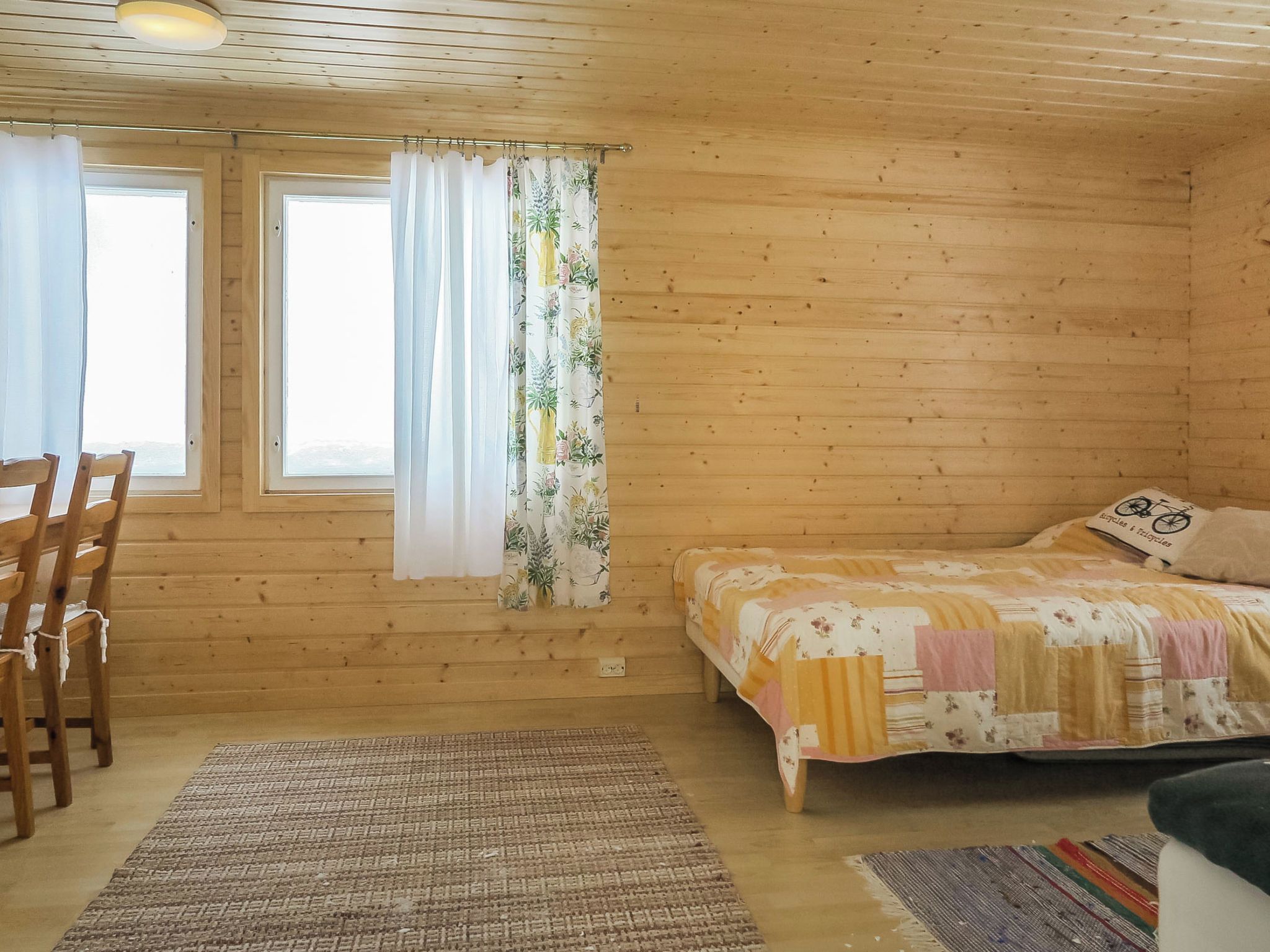 A wooden cabin bedroom with a bed, a chair, a window with curtains, and a view outside.