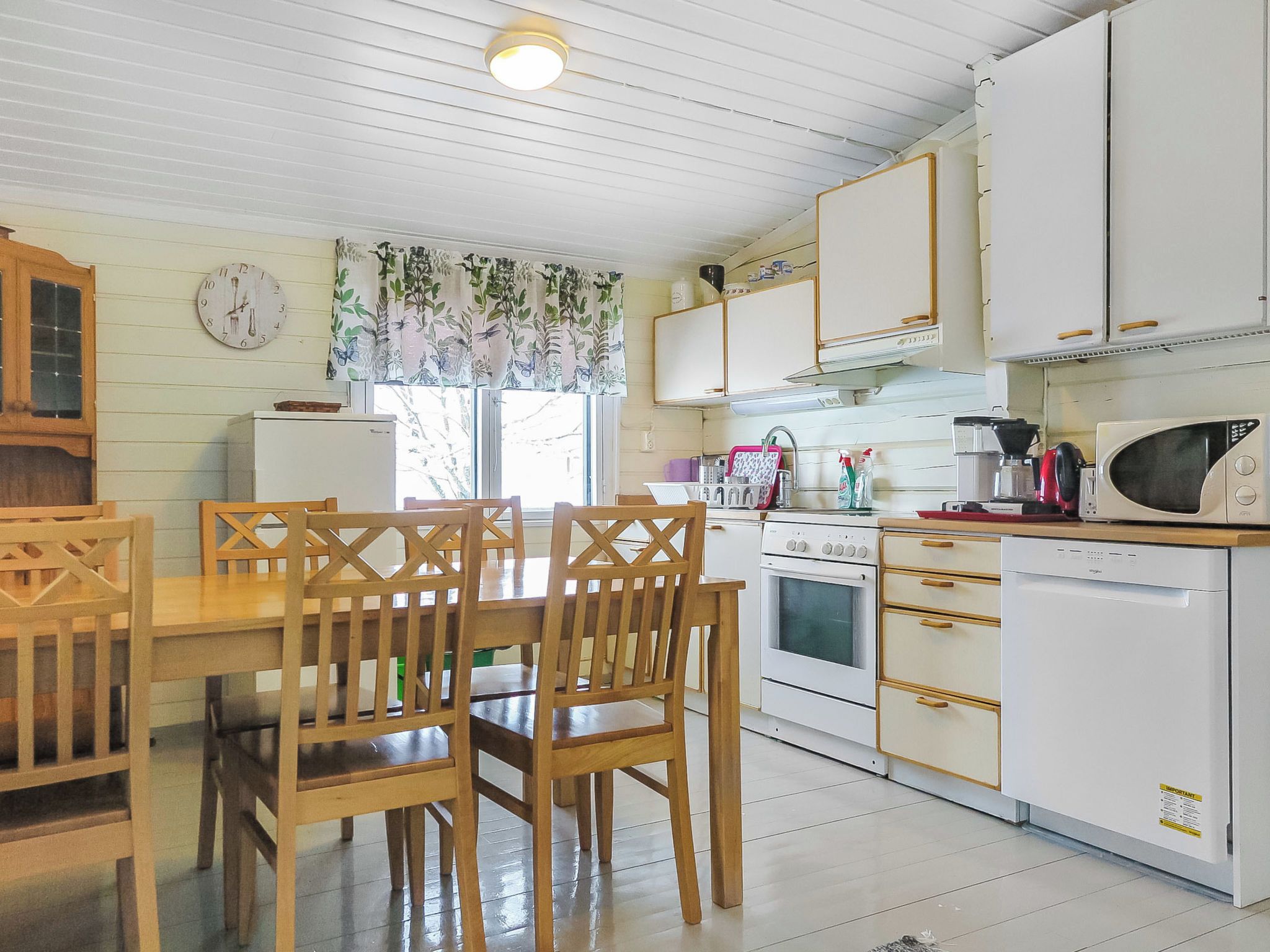 A compact kitchen with white cabinets, a wooden dining table with chairs, a refrigerator, an oven, a microwave, and a window with floral curtains.