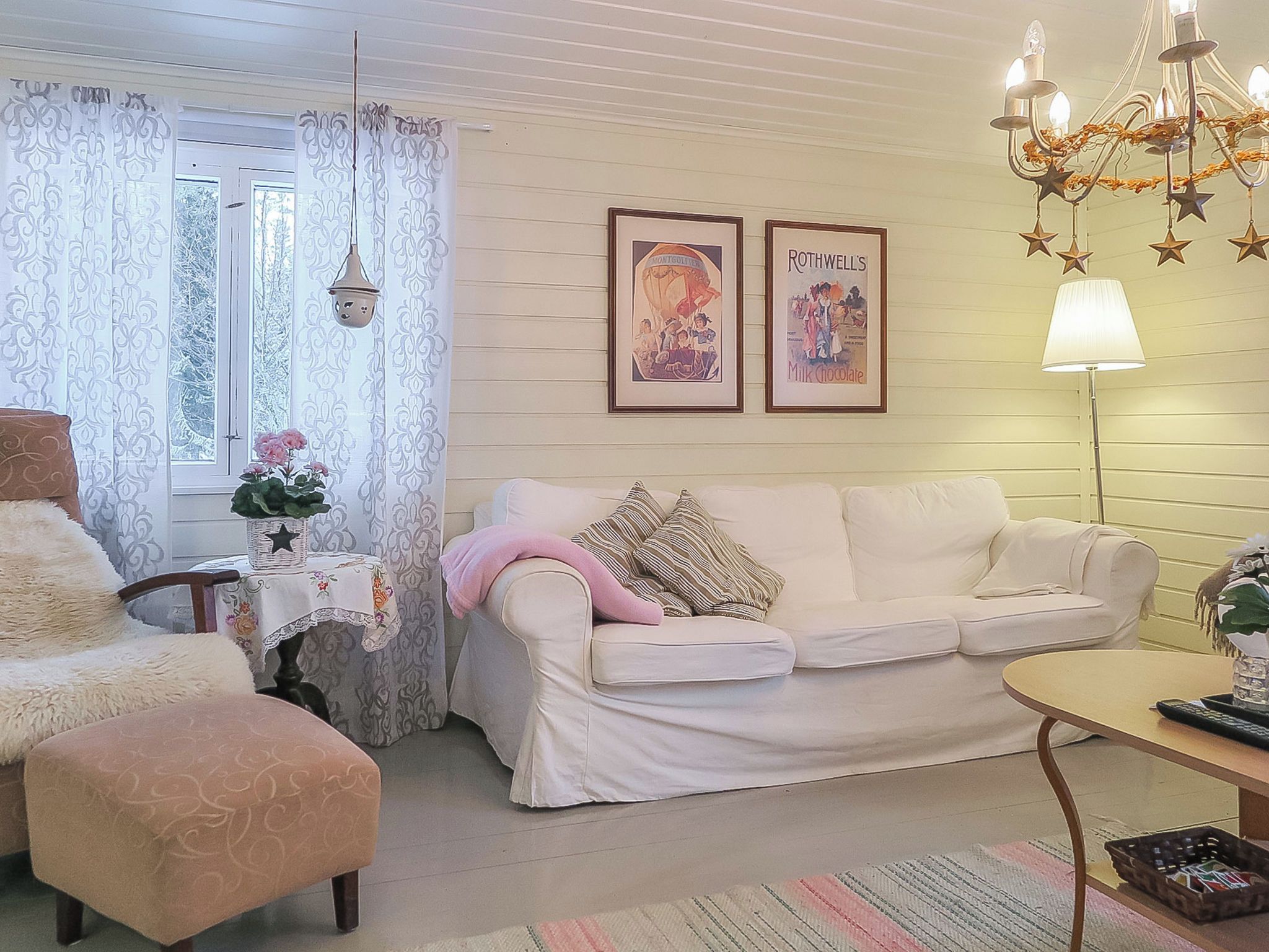 Una sala de estar acogedora con un sofá blanco, una mesita, una silla y una lámpara. Hay dos cuadros en la pared.