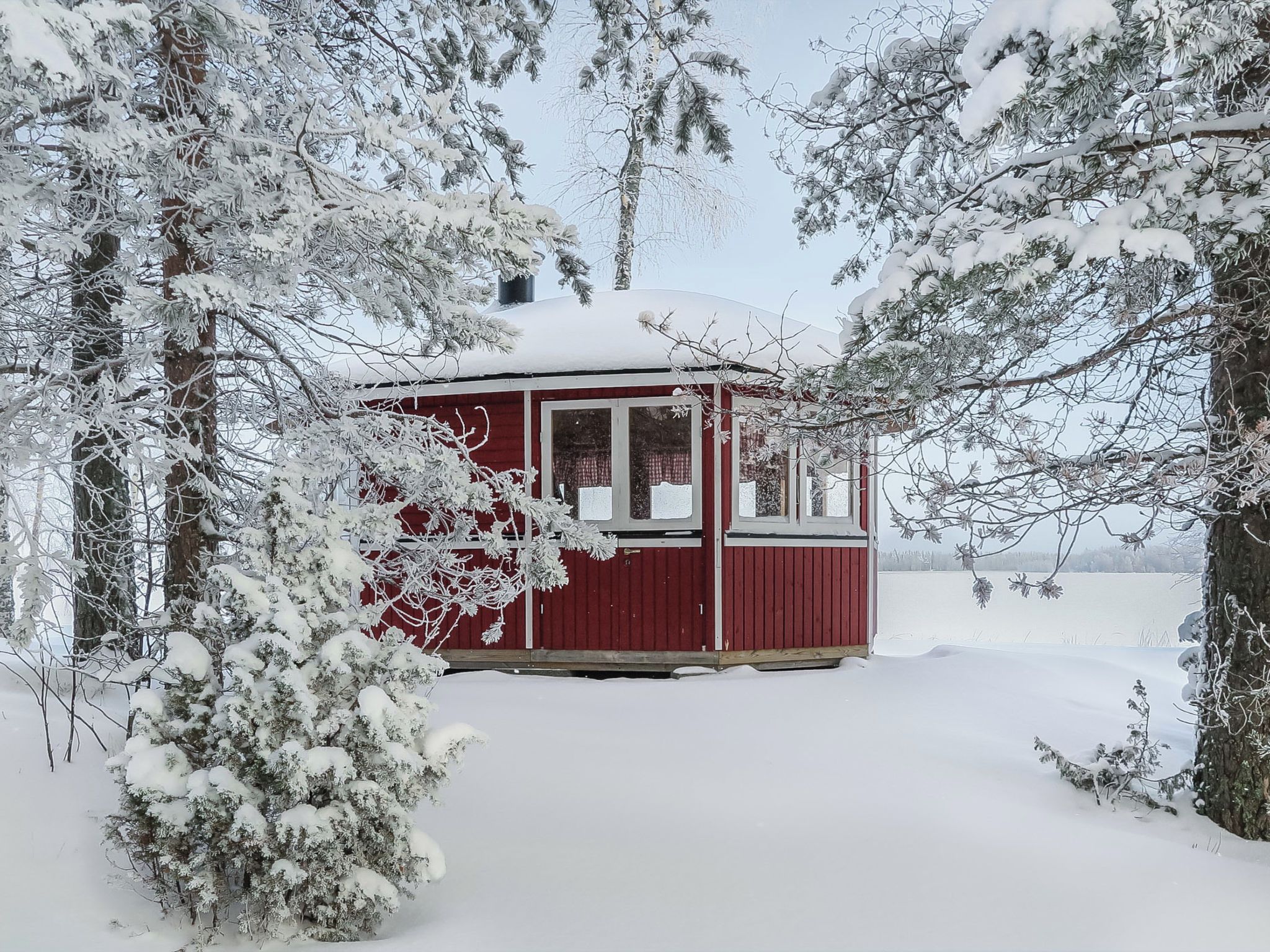 Lumipeitteinen mökki, jonka ulkoseinät ovat punaiset, ja jonka ympärillä on puita ja lumista maisemaa.