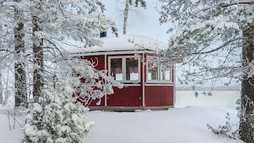 Lumipeitteinen mökki, jonka ulkoseinät ovat punaiset, ja jonka ympärillä on puita ja lumista maisemaa.
