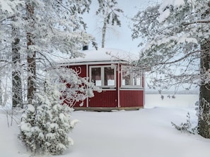 Eine mit Schnee bedeckte Holzhütte mit rotem Anstrich, umgeben von Bäumen und verschneiter Landschaft.