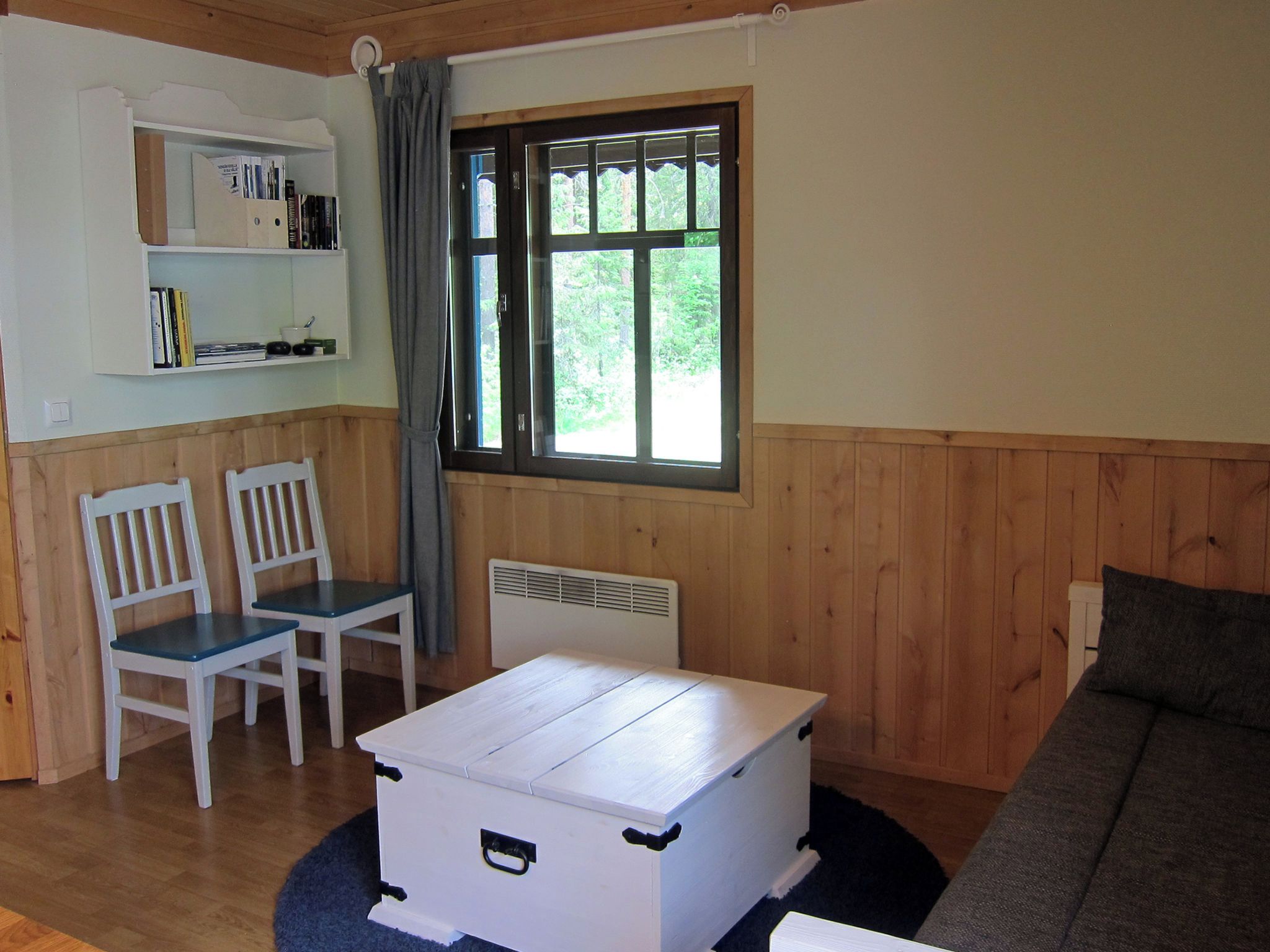 Une pièce avec une commode blanche, deux chaises blanches, un canapé gris, une fenêtre donnant sur un paysage verdoyant et une bibliothèque remplie de livres et surmontée d’une tasse.