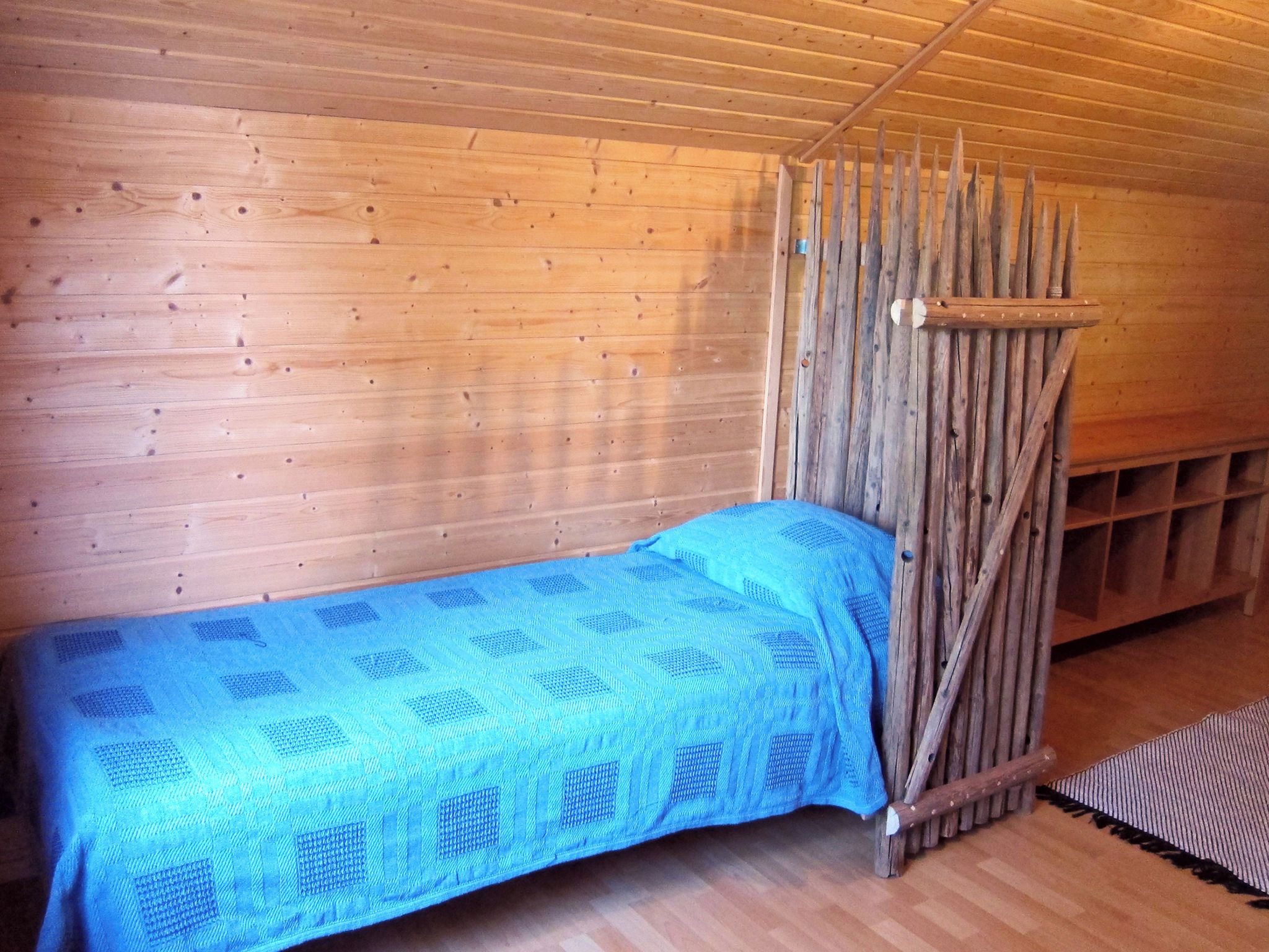 Un lit simple avec une couverture bleue, dans une pièce en bois dont le plafond est incliné.