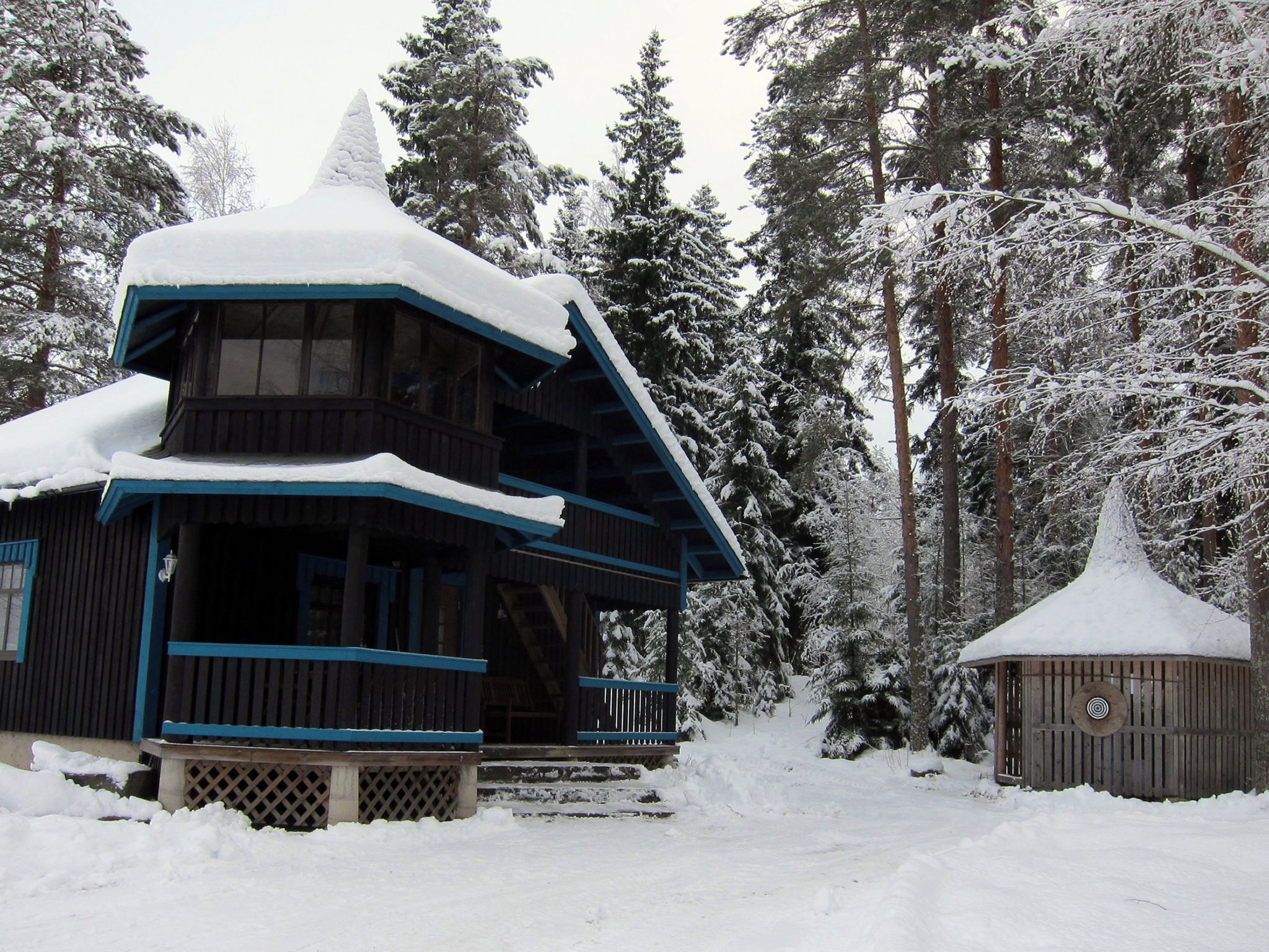 Una cabaña nevada con una chimenea destacada, rodeada de altos árboles de hoja perenne.