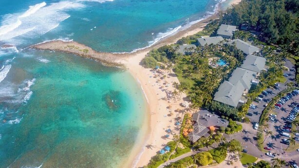 Luftaufnahme eines Küstenresorts mit Strand, klarem Wasser und einem Parkplatz.