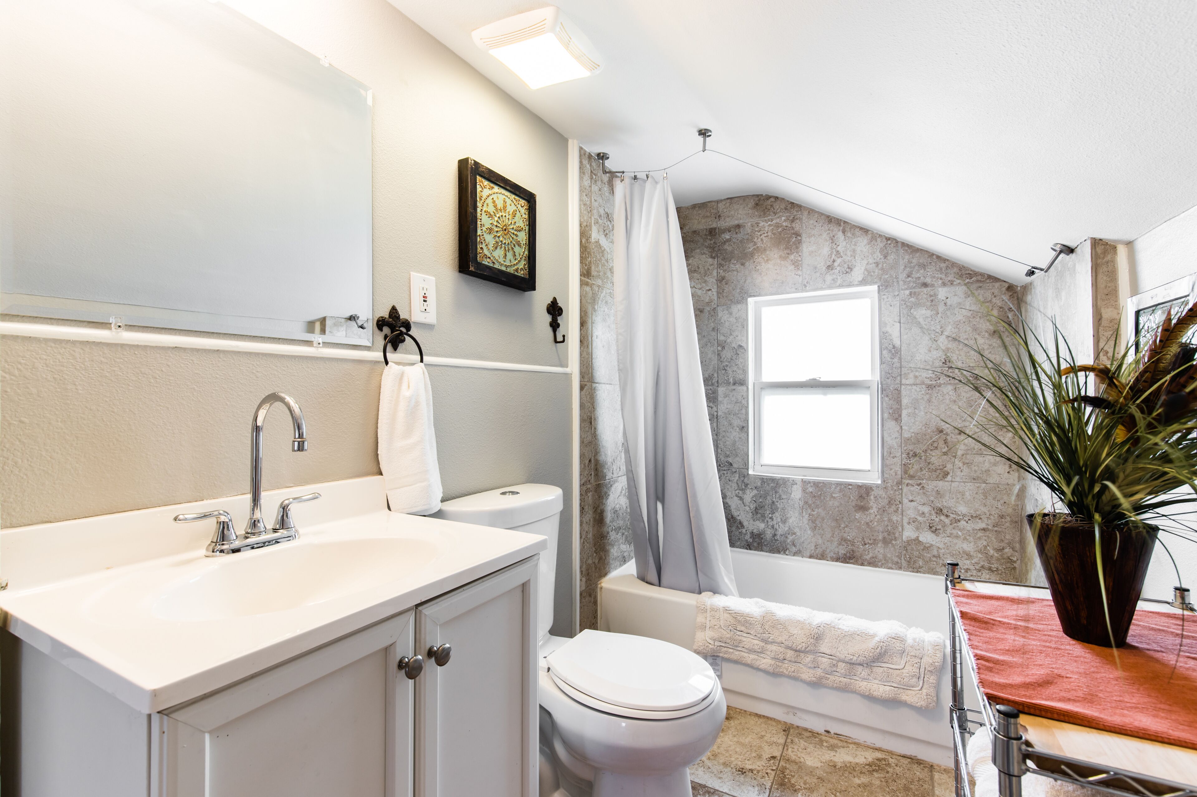 A bathroom with a white sink, toilet, and bathtub. There is a window with a view of the outside, and a plant on the bathtub's edge.