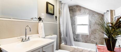 A bathroom with a white sink, toilet, and bathtub. There is a window with a view of the outside, and a plant on the bathtub's edge.