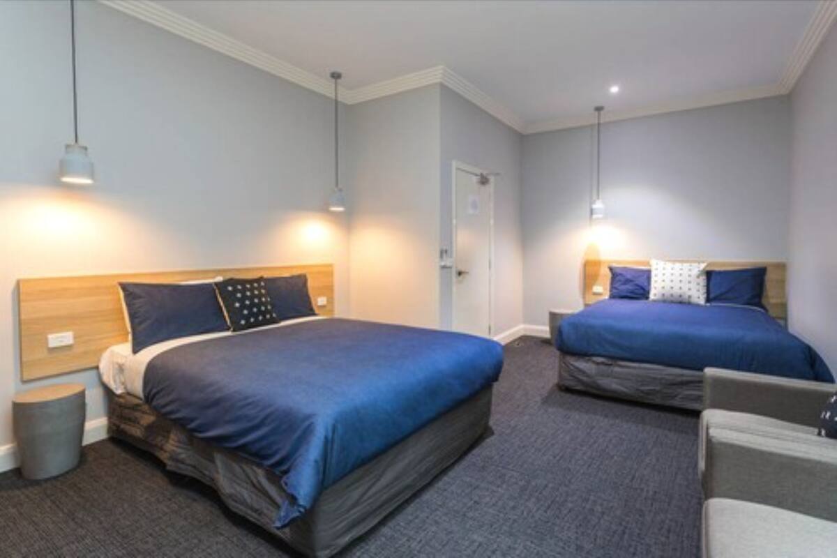 A hotel room with two beds, each with blue bedding and headboards, a small bedside table, a waste bin, and a door.