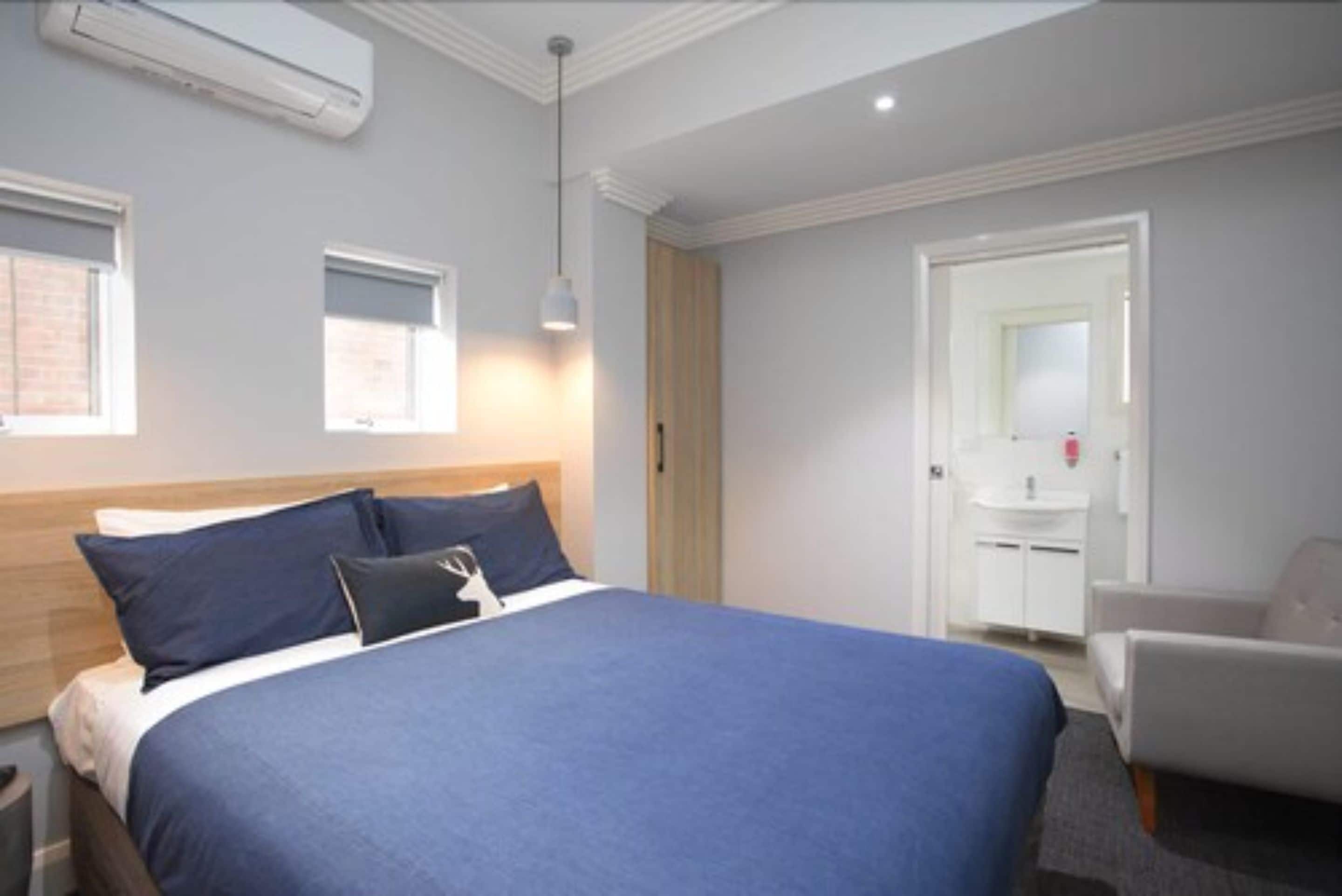 Une chambre avec un grand lit, une table de chevet, une chaise, une armoire, une fenêtre et une salle de bain avec un lavabo et un miroir.