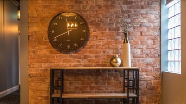 Un mur de briques avec une horloge murale ronde et une étagère ornée d’objets décoratifs.