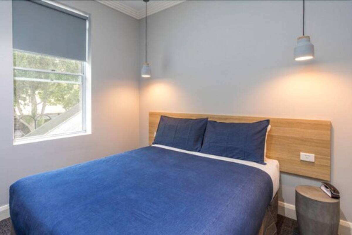 A neatly made bed with a blue bedspread and two pillows, a wooden headboard, a bedside table with a lamp, and a window with a view of trees.