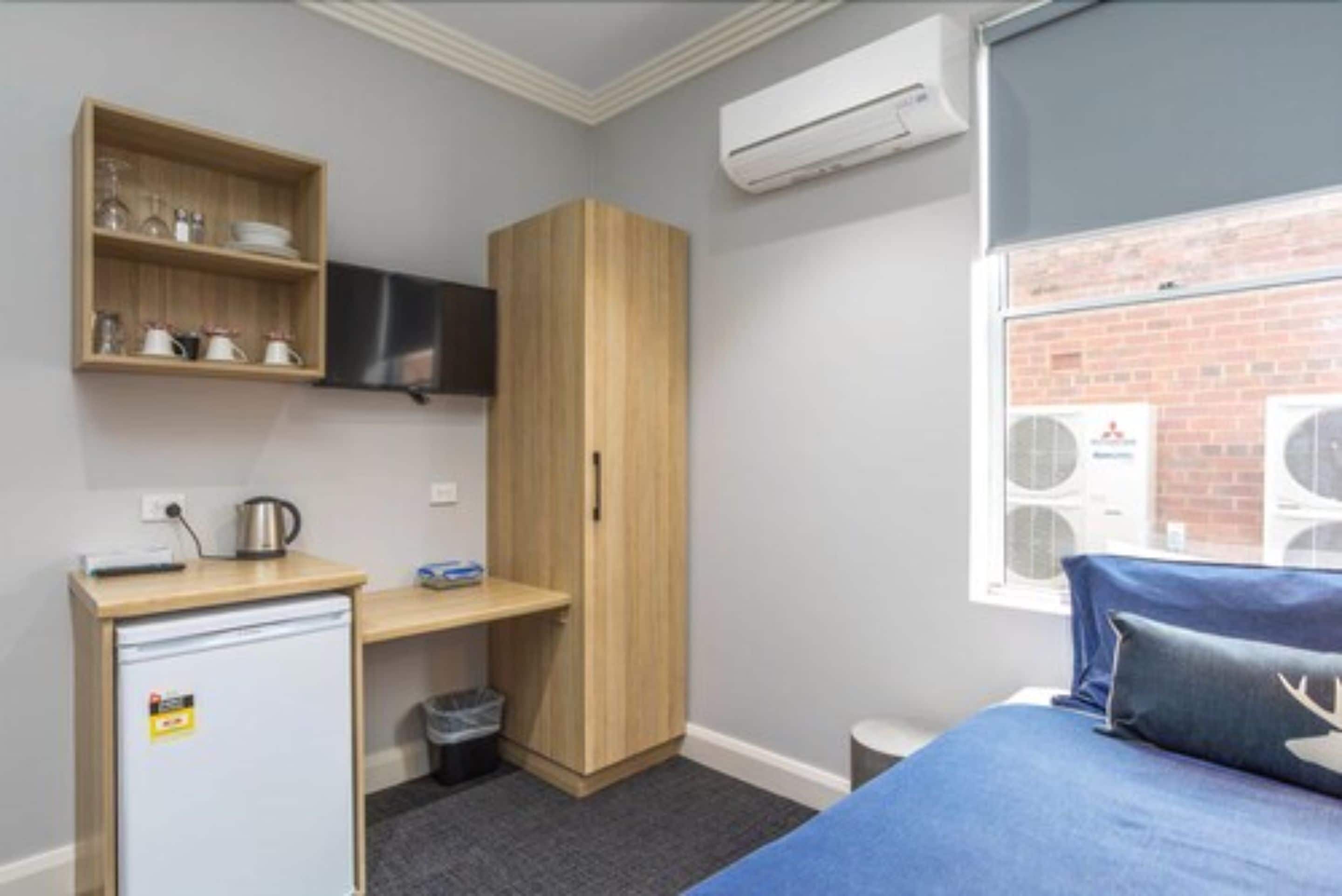Ein kleines Zimmer mit einem Bett, einer kleinen Küchenzeile, einem Holzschrank, einem an der Wand montierten Fernseher und einem Fenster mit Klimaanlagen.