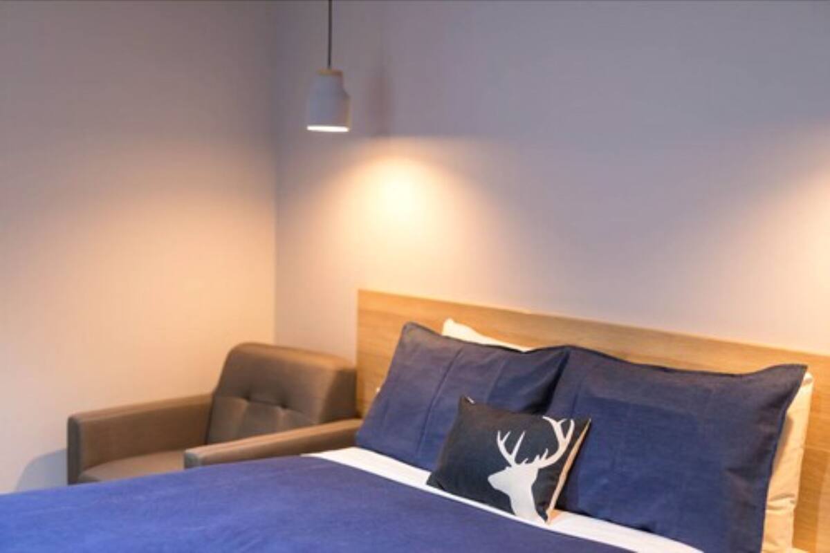 A neatly made bed with a blue bedspread and two pillows, one with a deer head design. There is a wooden headboard, a bedside table with a lamp, and a chair to the left.