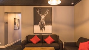 Un salon moderne avec un canapé foncé, deux coussins rouges et une table basse en bois. Un grand tableau représentant un cerf orne le mur.