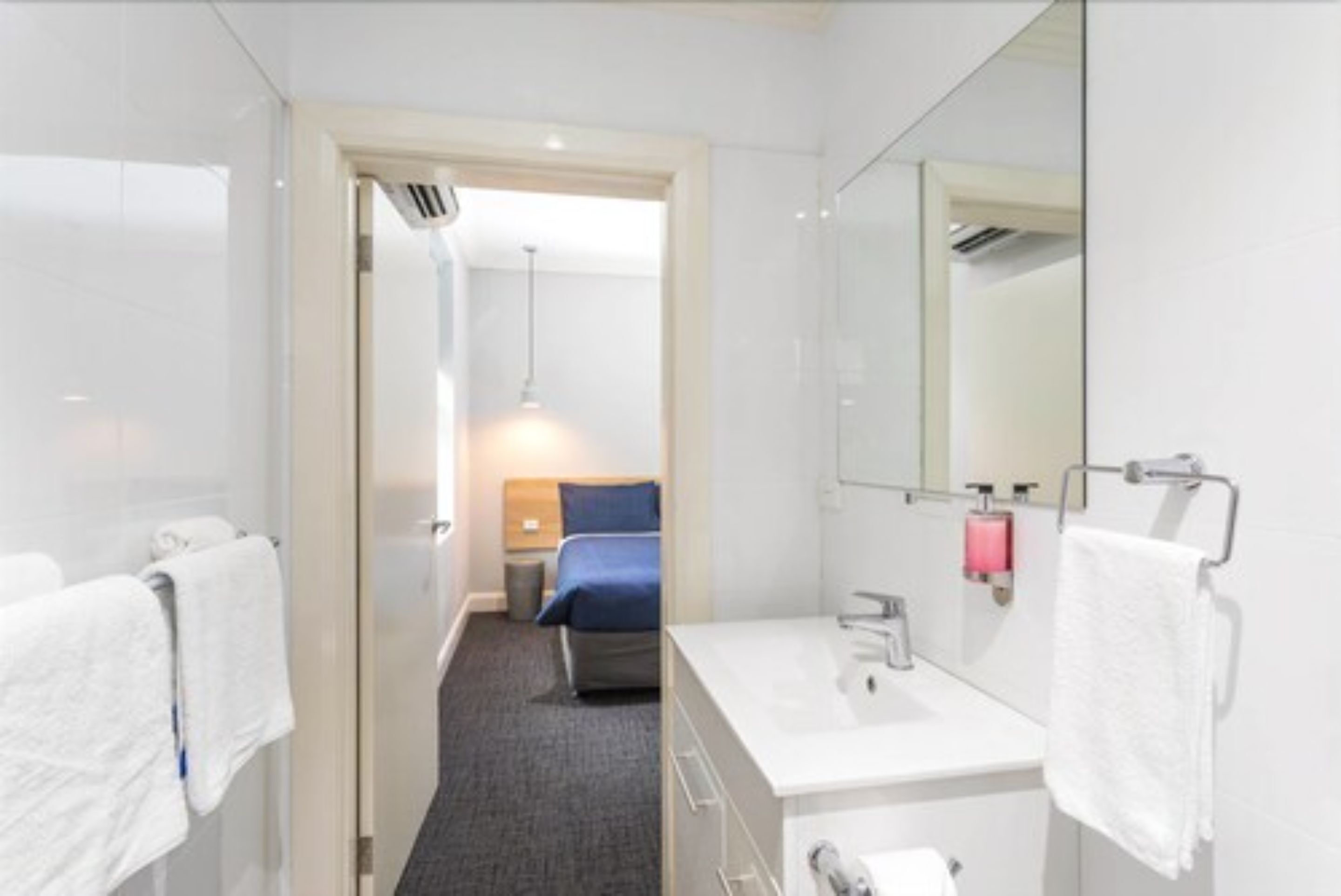 Moderni hotellihuone, jossa on kylpyhuone, sinisellä peitolla varustettu sänky ja näkyviin jäävä käytävä, joka johtaa toiseen huoneeseen.