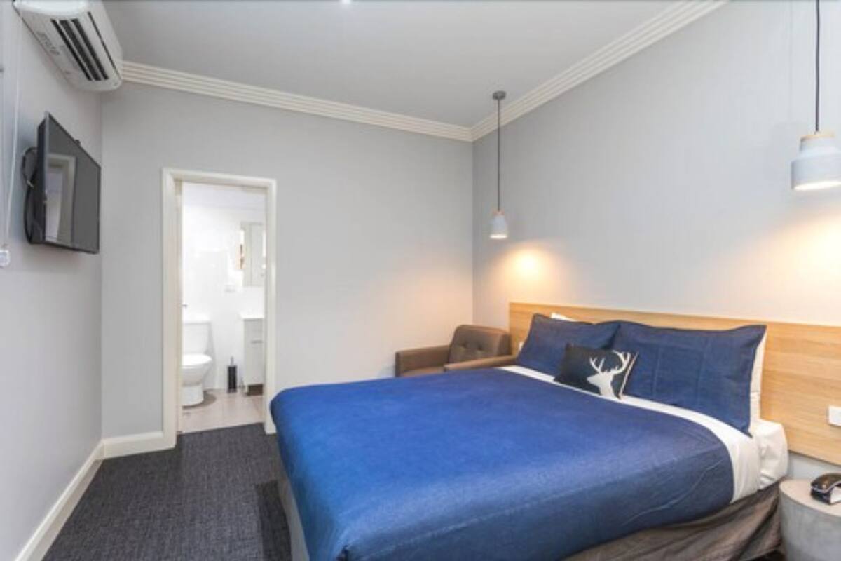 A hotel room with a large bed, a chair, a bedside table, a TV, and an air conditioning unit.