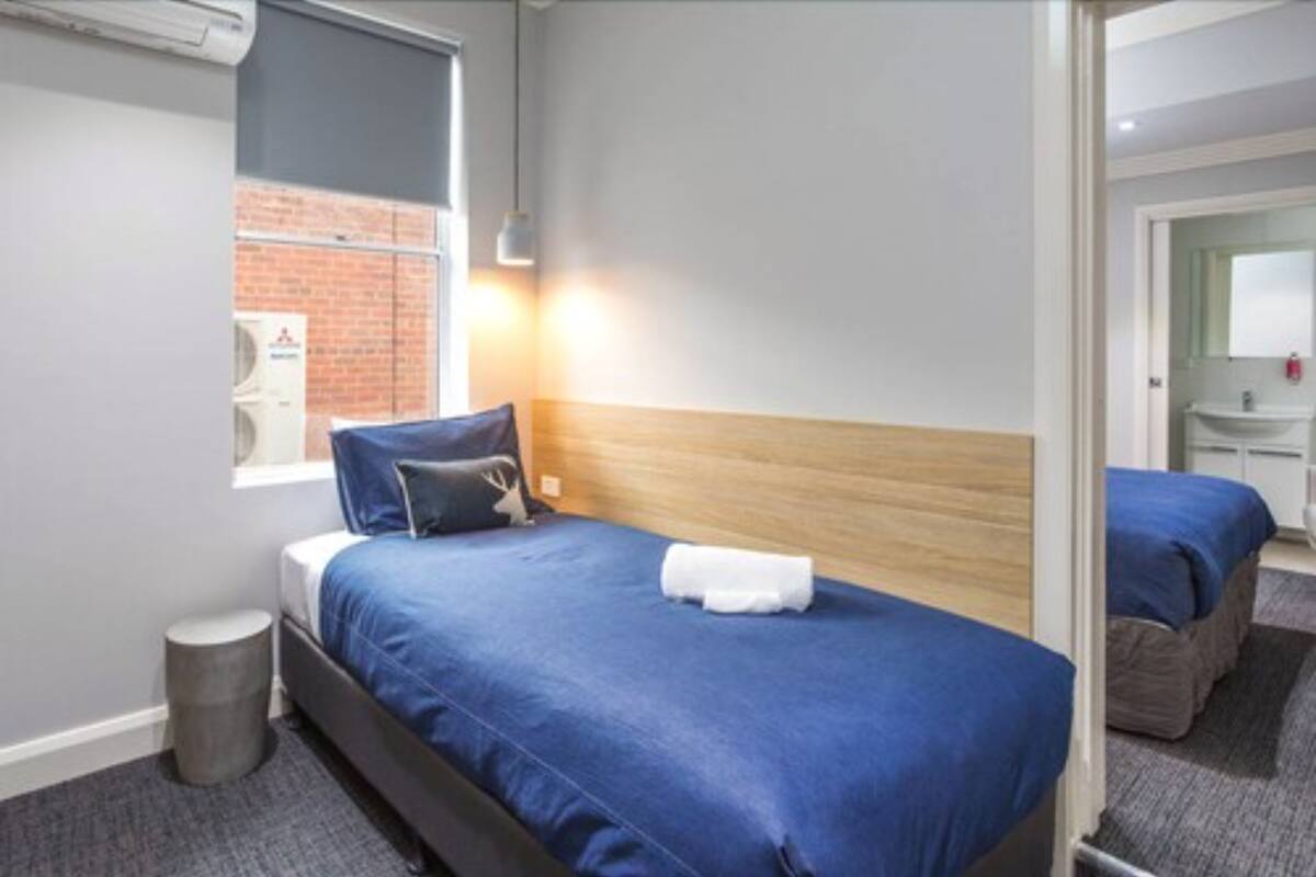 A hotel room with a bed, a small round stool, a window with a view of a brick wall, and a bathroom visible through an open door.