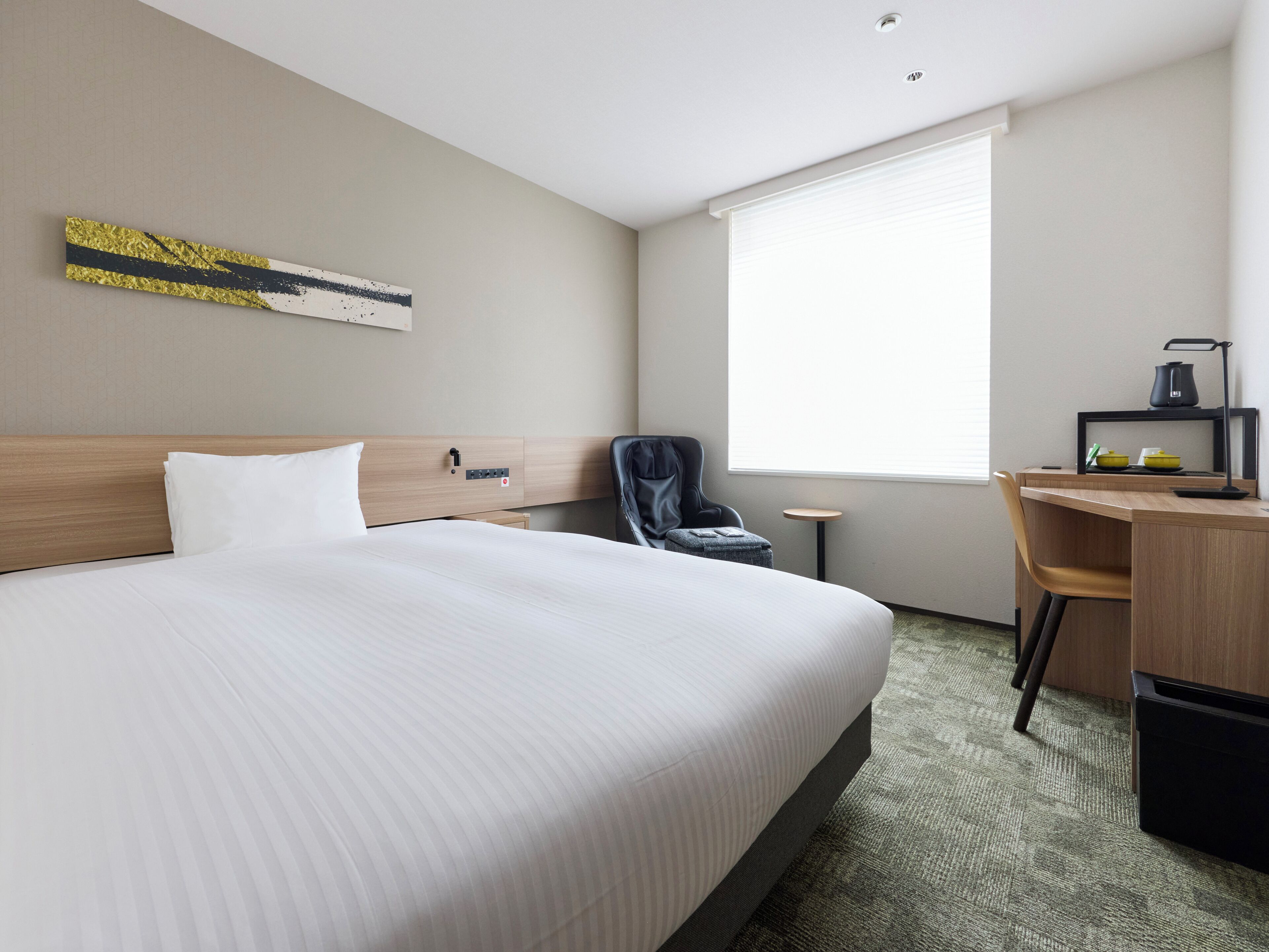A hotel room with a large bed, a desk with a chair, a small table, a chair, a window, and a wall-mounted artwork.