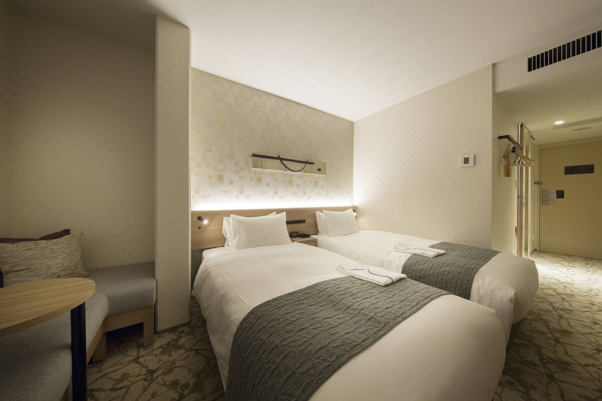 A hotel room with two beds, a seating area, a shower, and a wall-mounted light fixture.