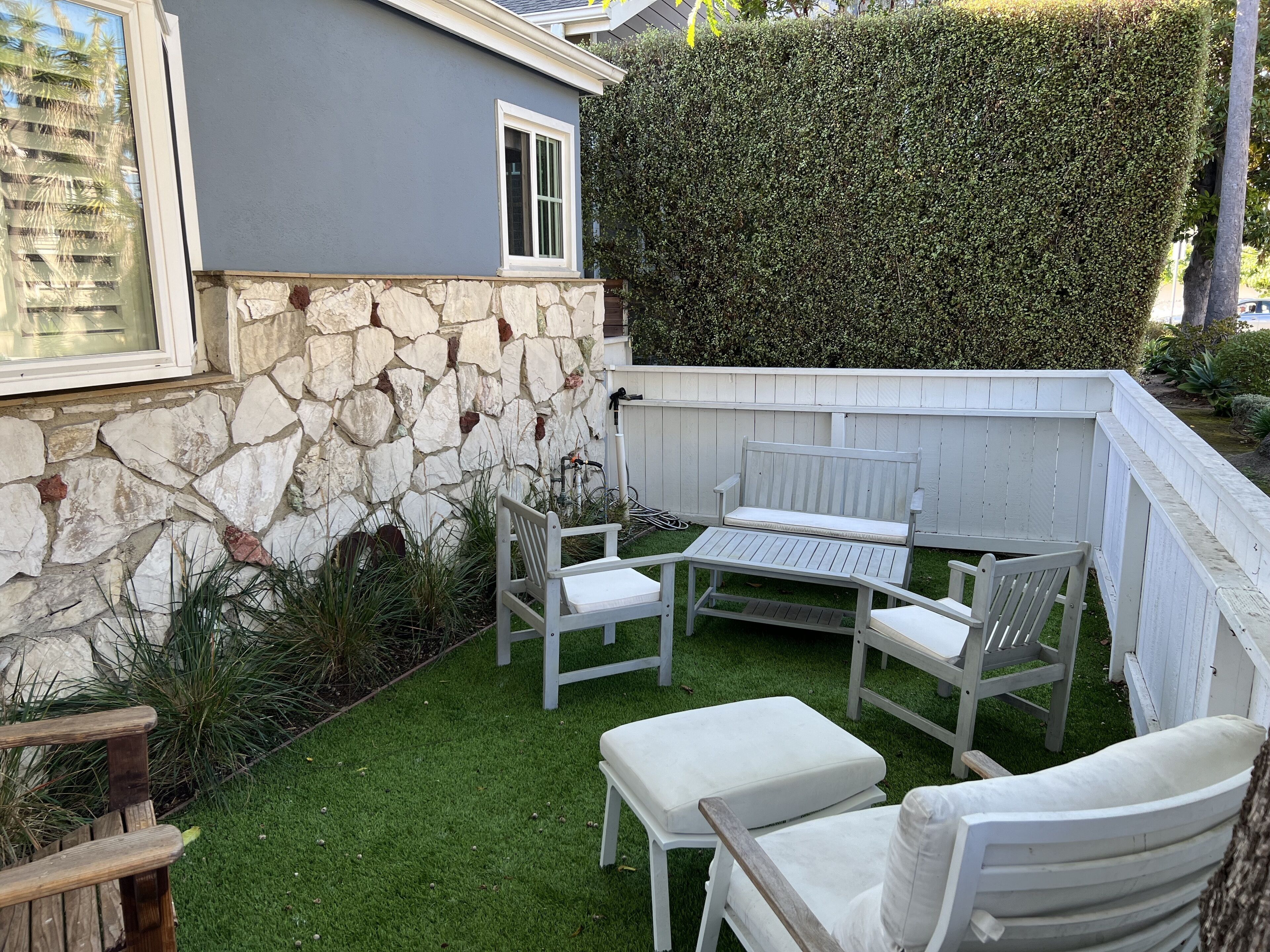 Un patio con pared de piedra, mobiliario de madera blanca y seto.