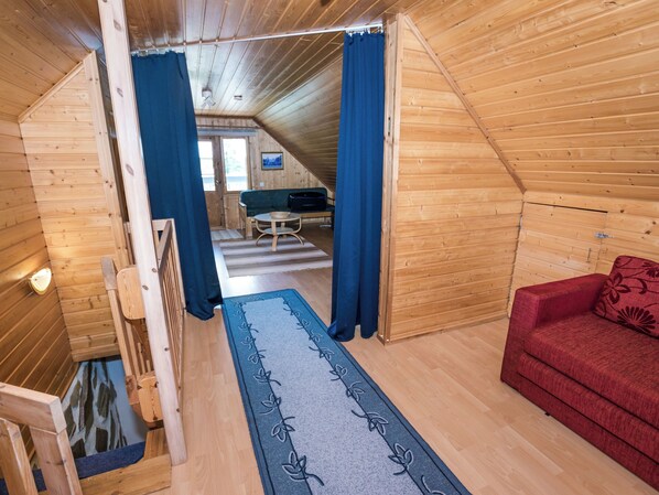 Interior de una cabaña de madera con un sofá, una silla roja, una cortina azul y una escalera de madera.