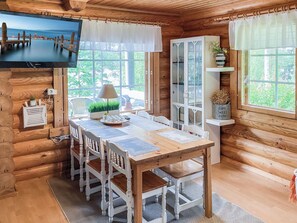 A wooden cabin interior with a dining table, chairs, a television mounted on the wall, and large windows with curtains.