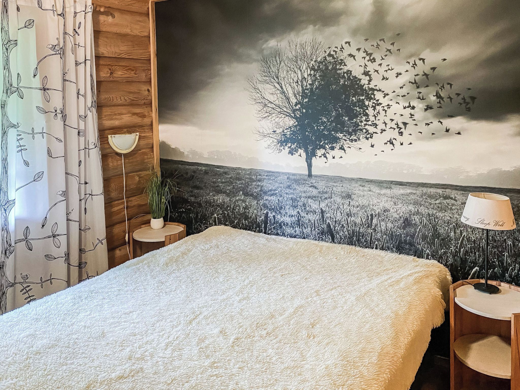 A bedroom with a bed, a nightstand, a lamp, a potted plant, and a wall mural of a tree and birds.