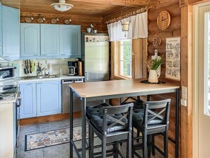 A kitchen with light blue cabinets, a wooden table, and two chairs. There is a refrigerator, a coffee maker, and a window with curtains.