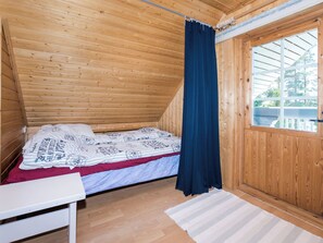 Une petite cabane d’une seule pièce, avec un plafond en pente, un lit recouvert d’une literie à motifs, une table blanche et une fenêtre avec un rideau.