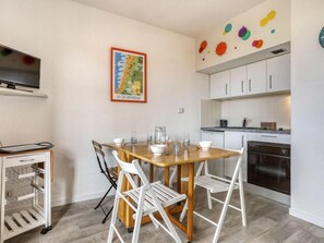 A compact kitchen with white cabinets, a wooden dining table with four chairs, and a small TV mounted on the wall.