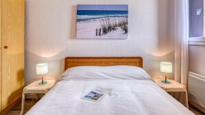 A bedroom with a bed, two bedside tables with lamps, a wooden wardrobe, a beach-themed painting on the wall, and a window with curtains. - Trinquart by Interhome (Seignosse)