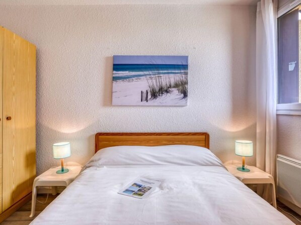 A bedroom with a bed, two bedside tables with lamps, a wooden wardrobe, a beach-themed painting on the wall, and a window with curtains.
