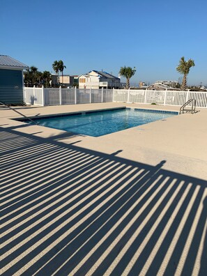 Una piscina con valla blanca, terraza con sombra y cielo azul despejado.