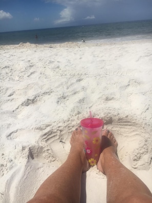 A sandy beach with a clear view of the ocean and a person relaxing with a drink.