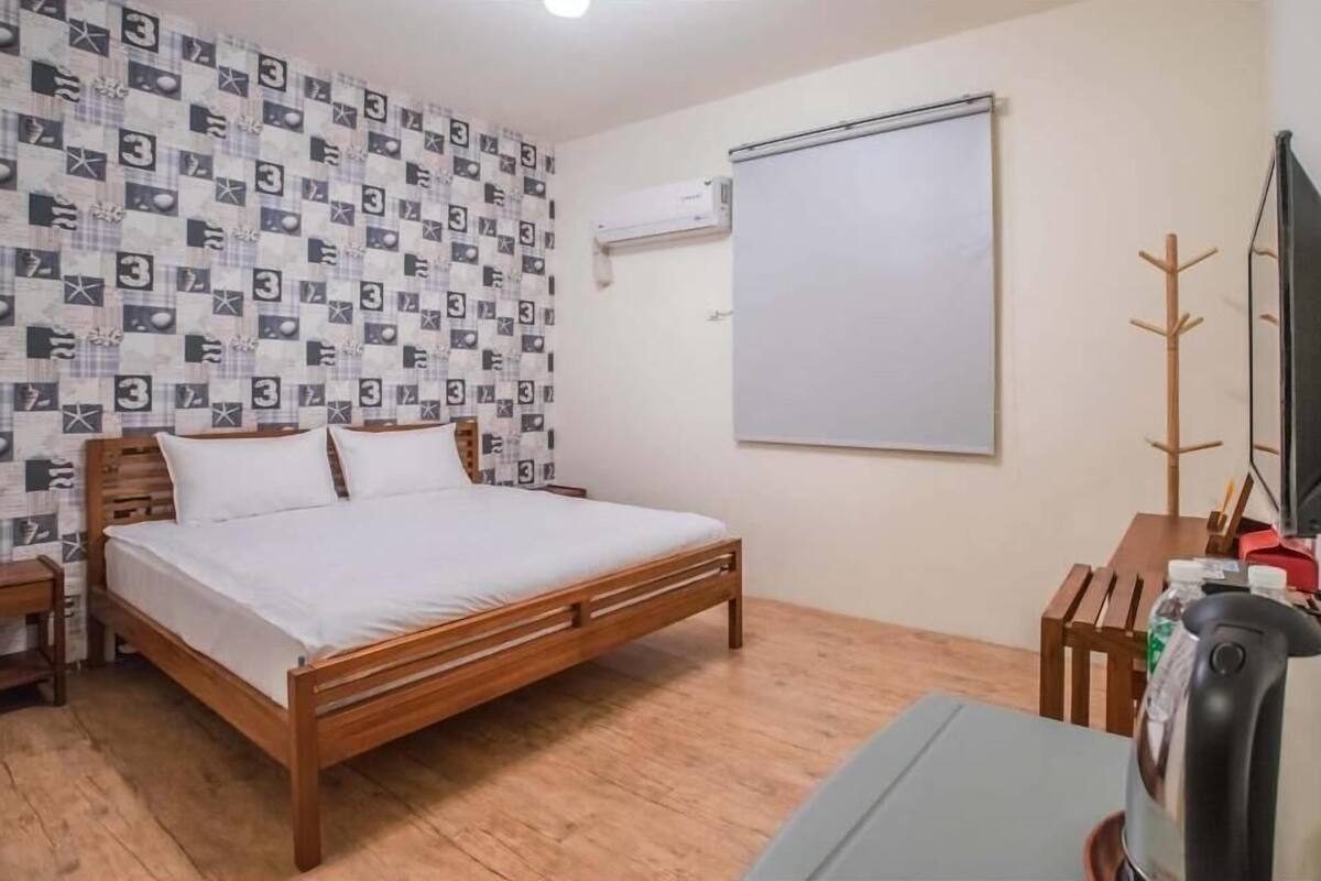 Une chambre avec un lit en bois, un mur blanc recouvert d’un papier peint à motifs, un tableau blanc, un porte-manteau et une bouilloire.