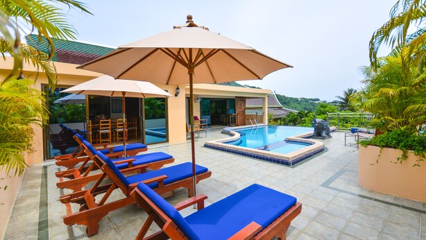 Un espace piscine avec des chaises longues et des parasols, une piscine et un bâtiment doté d’un toit en tuiles.