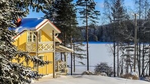 A yellow cabin with a red roof, situated by a frozen lake surrounded by trees.
