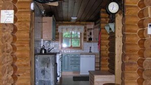 A rustic cabin interior with a wooden log wall, a kitchen area with a sink, cabinets, and a window with curtains.