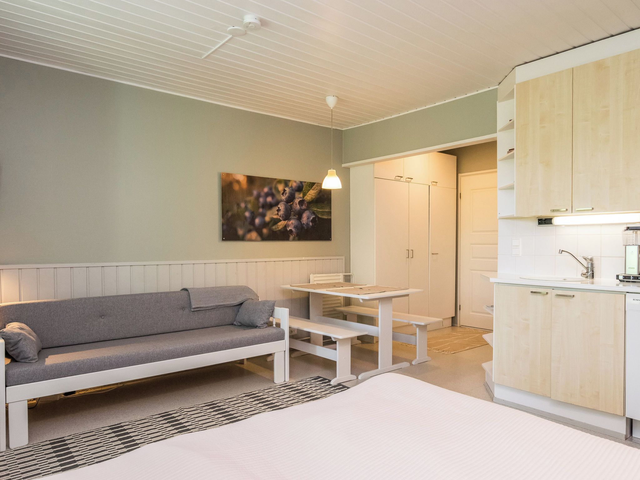 Habitación compacta con sofá, una mesita, un banco, zona de cocina con armarios y una lámpara en el techo.