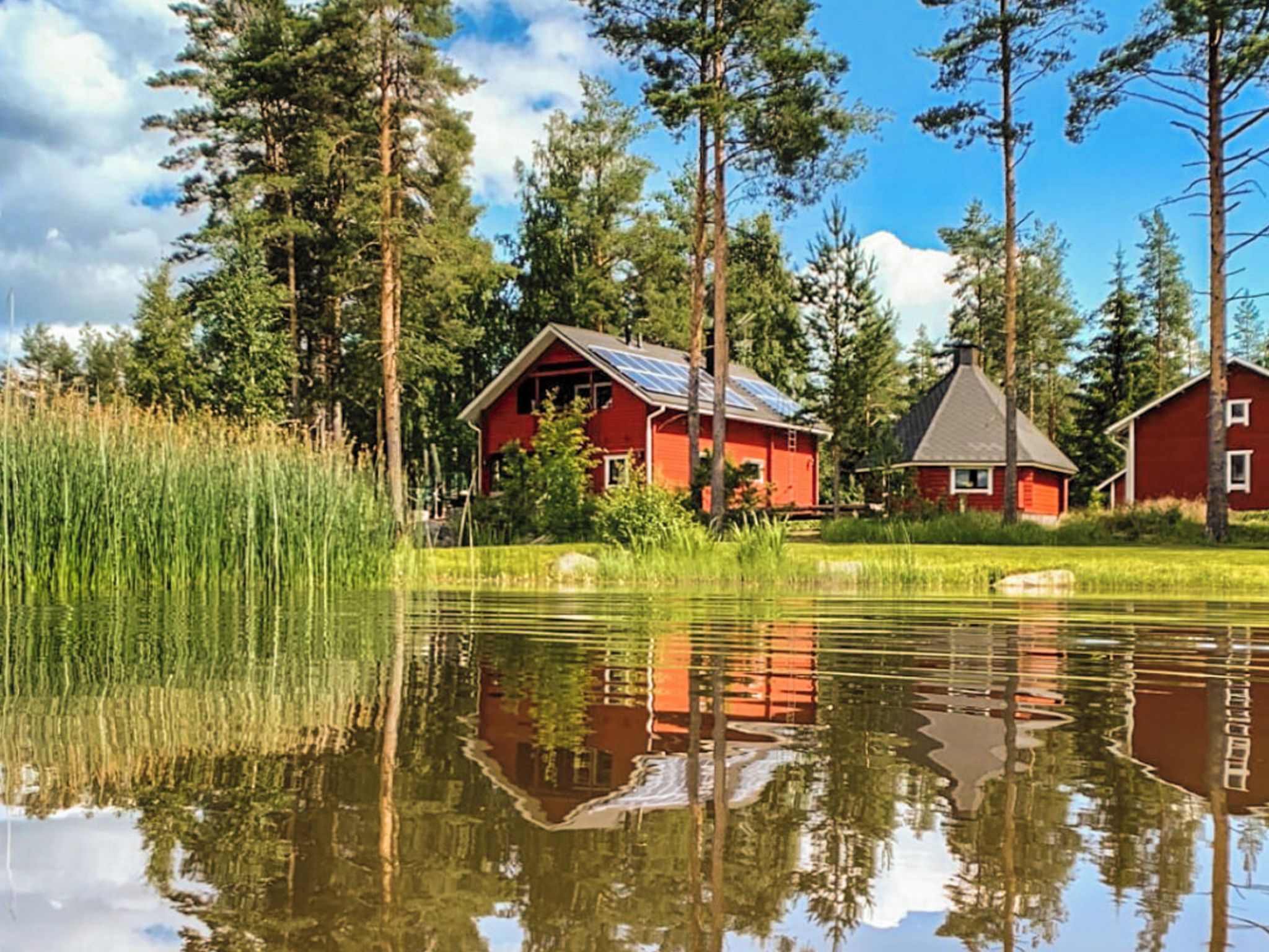 En rød hytte med solpaneler på taget, omgivet af høje træer og et roligt vandareal der spejler omgivelserne.
