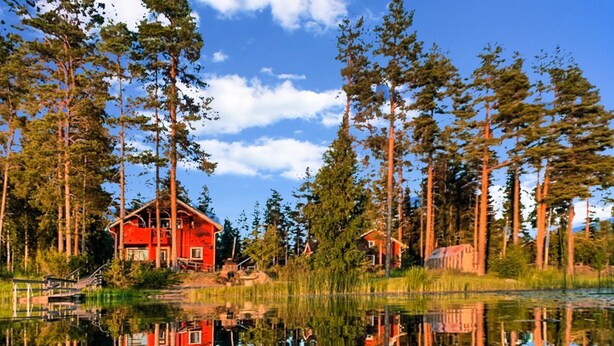 A red cabin surrounded by tall pine trees and a calm lake reflecting the scene. - Kallioniemi by Interhome (Kiuruvesi)