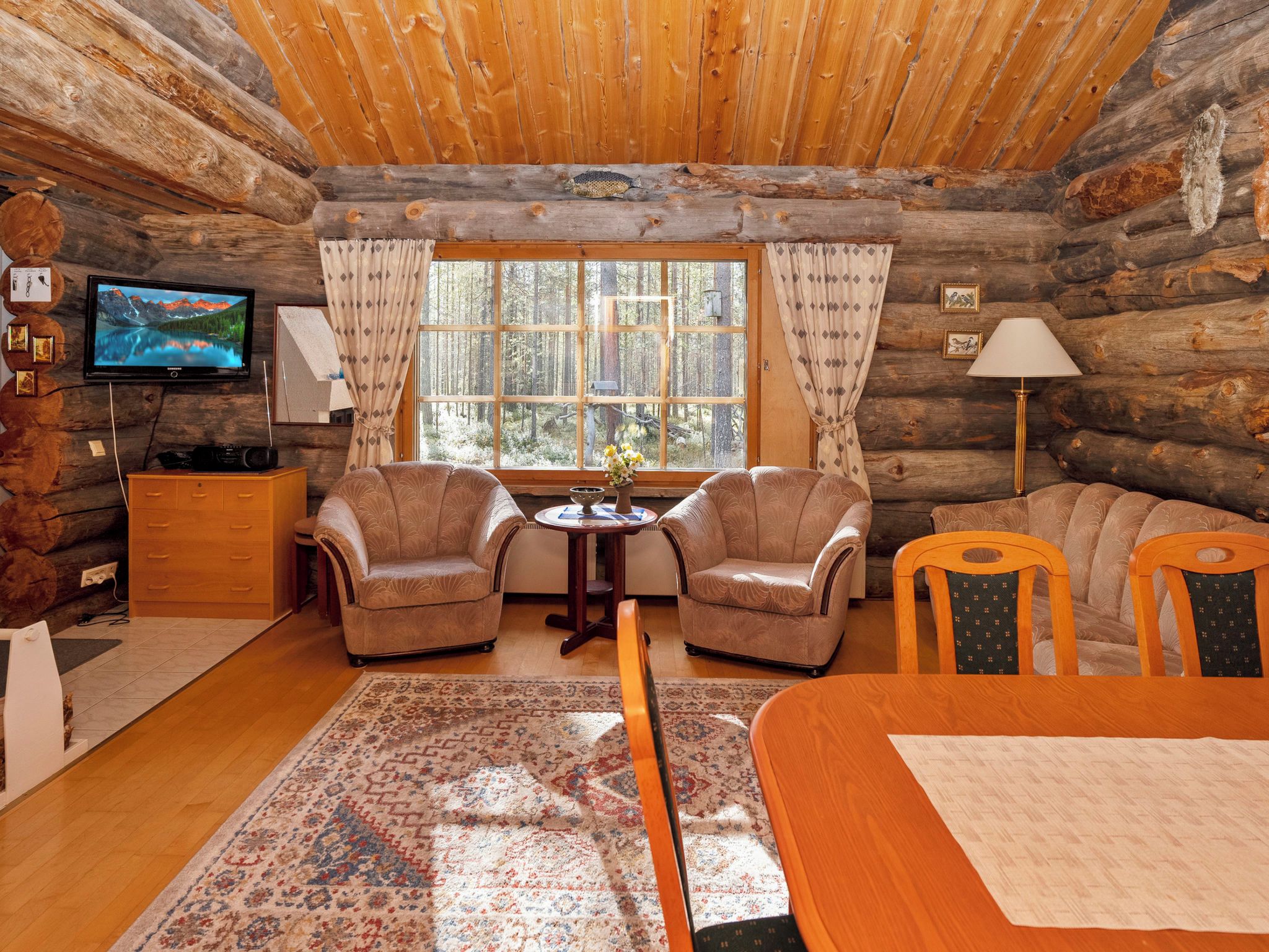 L’intérieur d’une cabane en rondins, avec un téléviseur, deux fauteuils, un canapé, une table à manger et une grande fenêtre avec des rideaux.