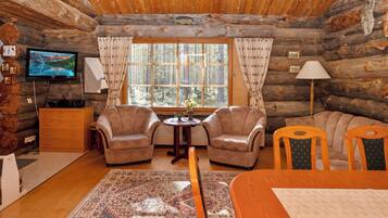 L’intérieur d’une cabane en rondins, avec un téléviseur, deux fauteuils, un canapé, une table à manger et une grande fenêtre avec des rideaux.